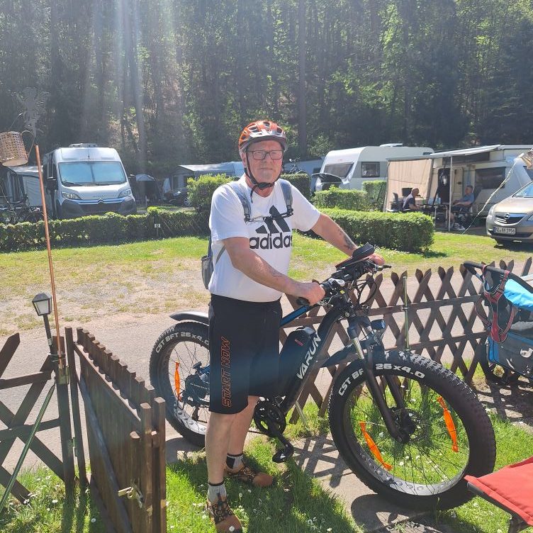 Mann mit Mountainbike in einem Campingbereich, umgeben von Wohnmobilen und Bäumen.